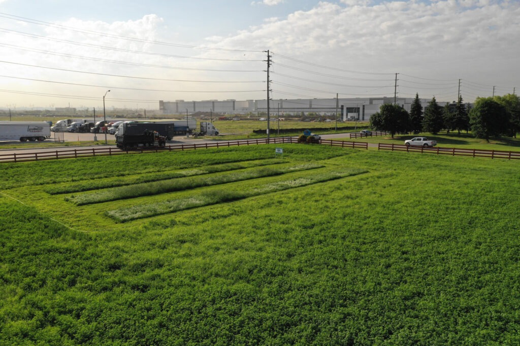 A green field with a parking area, trucks, electric poles, and a distant, large industrial building under a partly cloudy sky.