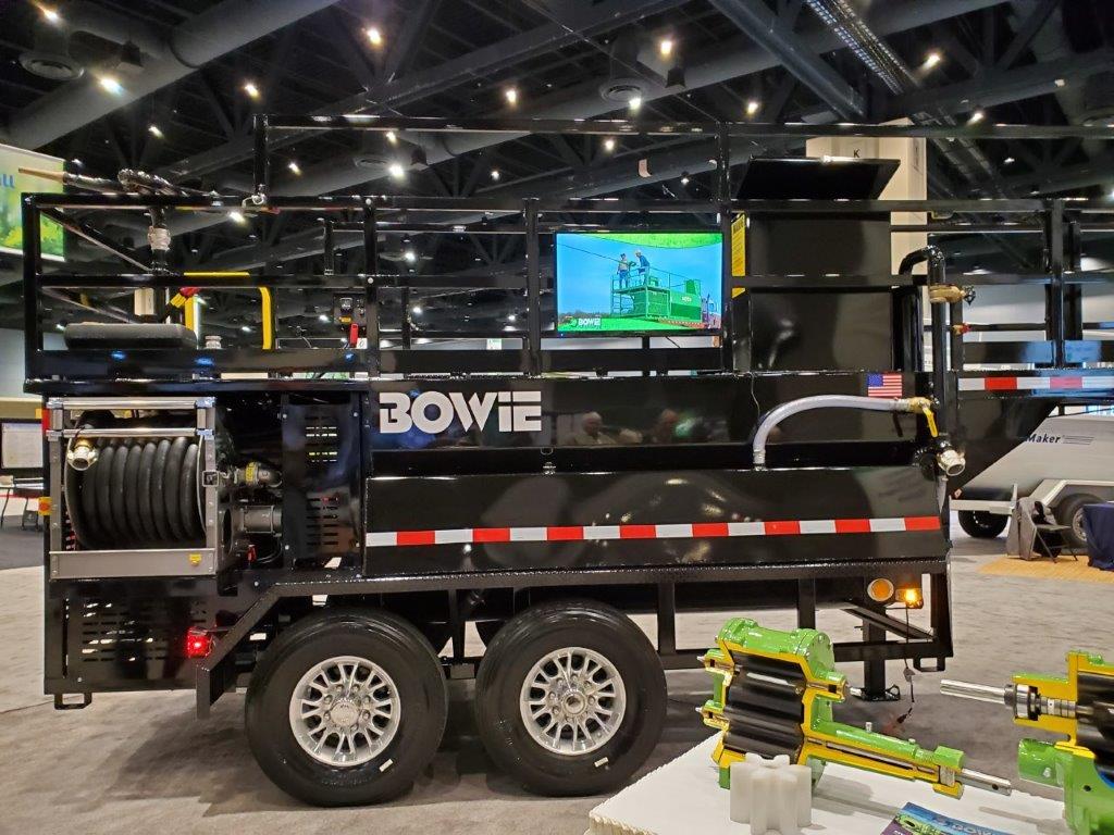 A black, heavy-duty trailer labelled "Bowie" is displayed indoors with industrial equipment around, and a screen showing related footage.