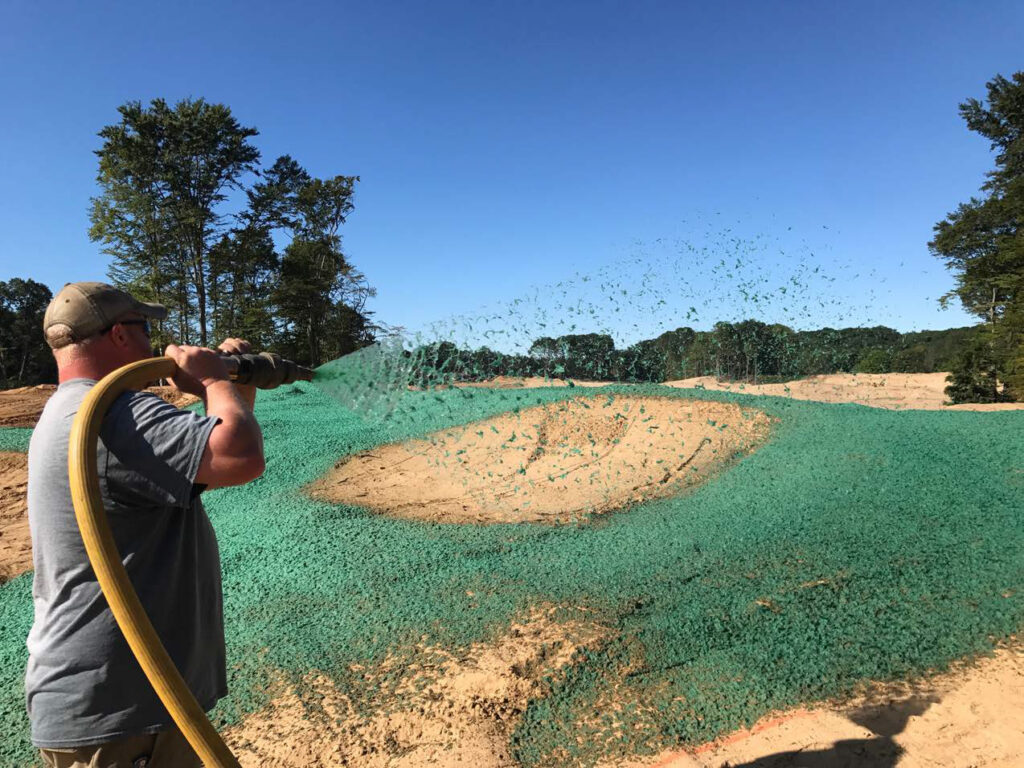 A person sprays green hydromulch on bare soil in an open field with trees in the background under a clear blue sky.
