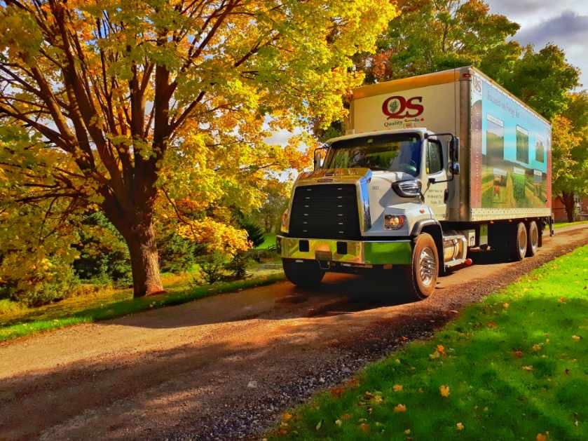 A large delivery truck drives down a tree-lined path with vibrant autumn foliage, casting shadows on the road. No landmarks or historical buildings present.