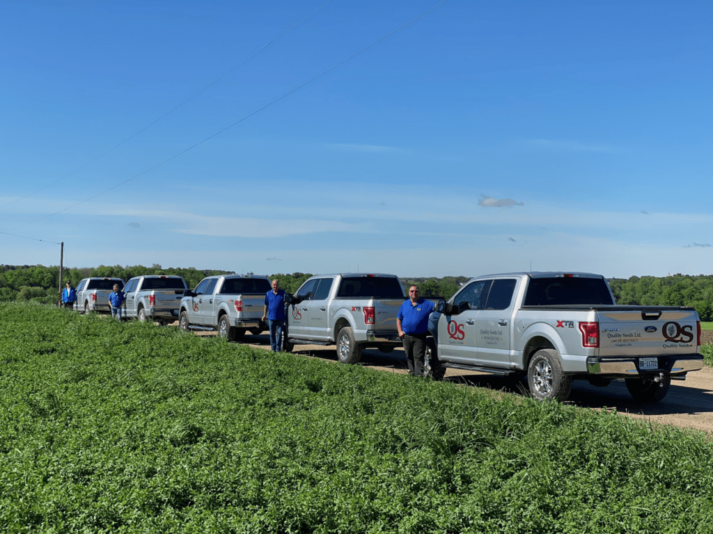 Several persons stand beside a line of Quality Seeds Ltd. pickup trucks on a rural road near a lush green field under a clear blue sky.