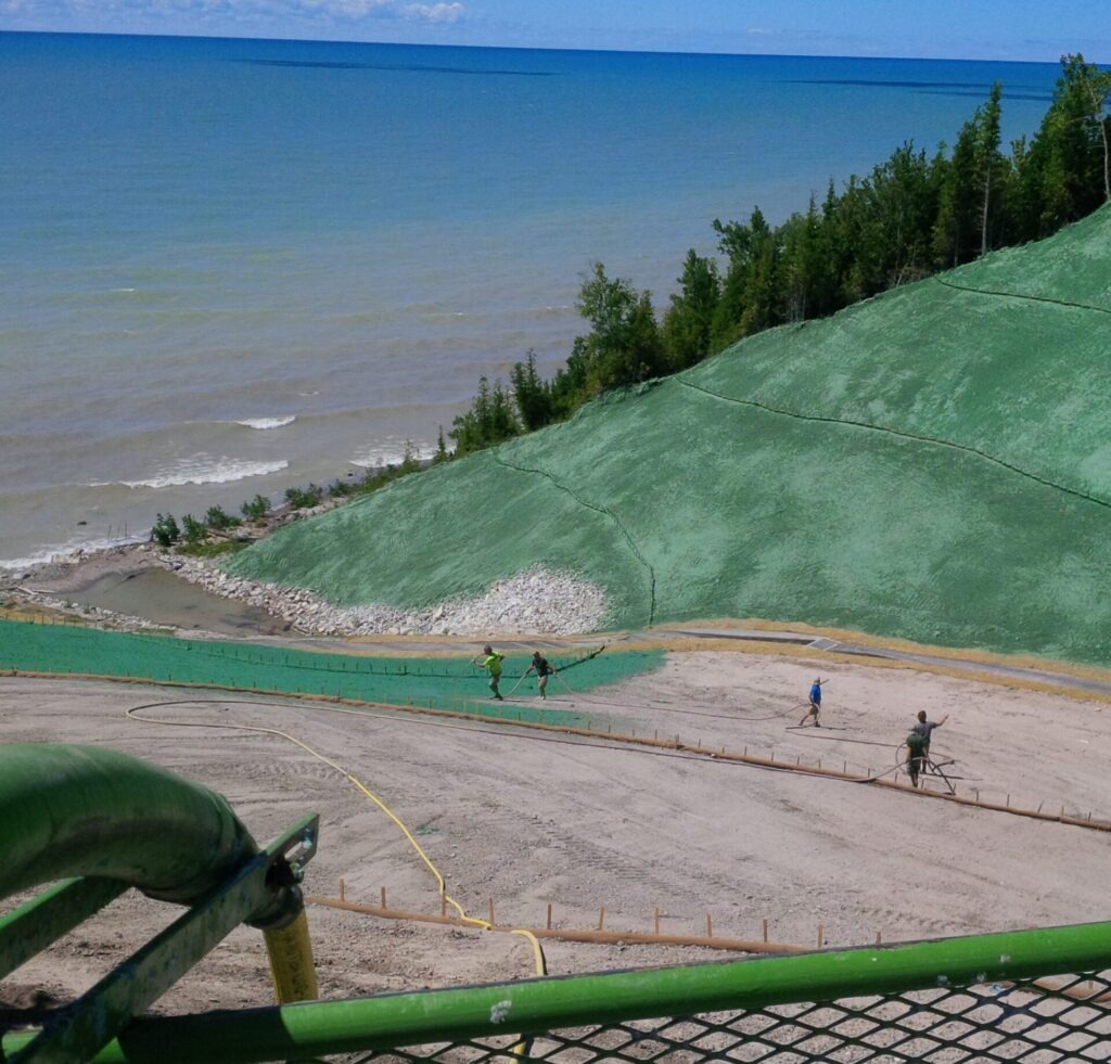 Four people work on a green slope near a lake, with a forested area in the background. The sky is blue with clouds.
