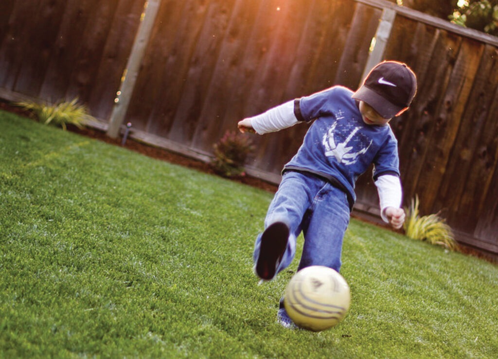 A child wearing a cap kicks a soccer ball on a grassy lawn, with a wooden fence and plants in the background under warm sunlight.