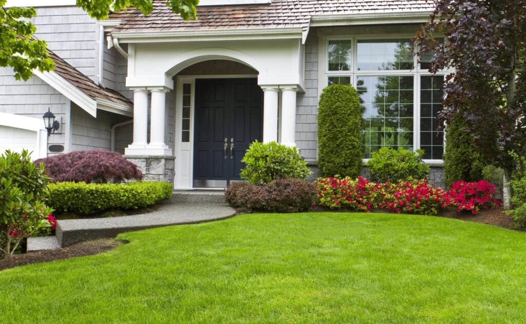 A well-maintained front yard with vibrant flowers and green grass leads to a house with a dark blue door and grayish exterior.