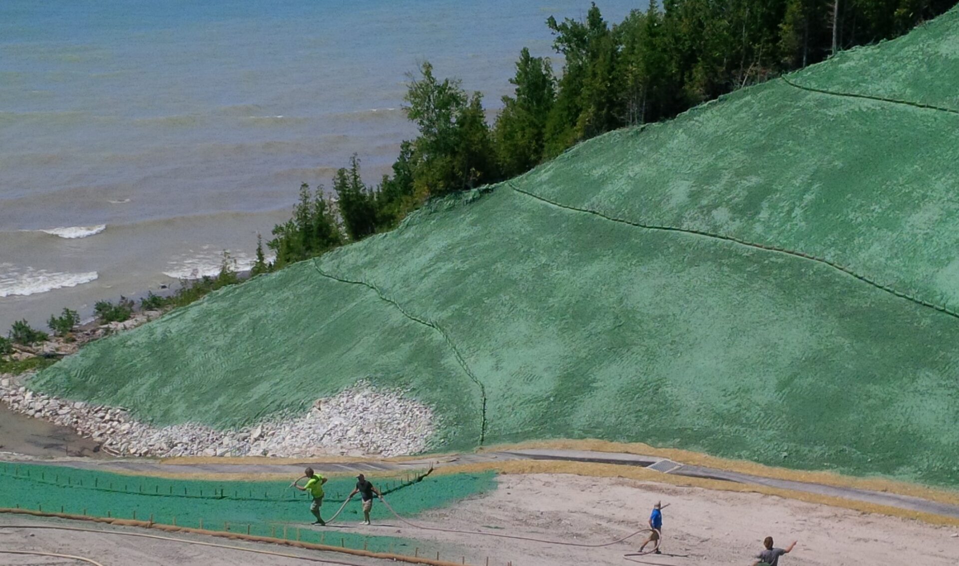 Three people work on hydroseeding a green hillside beside a lake, with trees in the background. The lake has gentle waves reaching the shore.