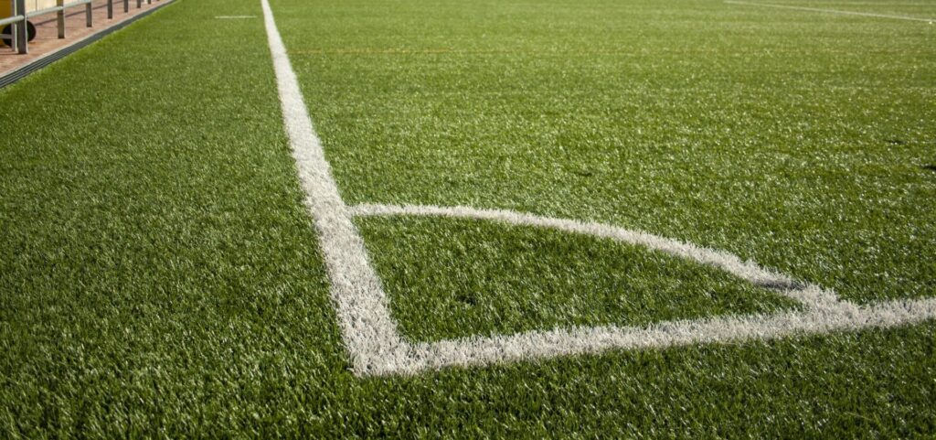 A green sports field featuring white boundary lines, with a stadium seating area and handrails visible along the top edge of the image.